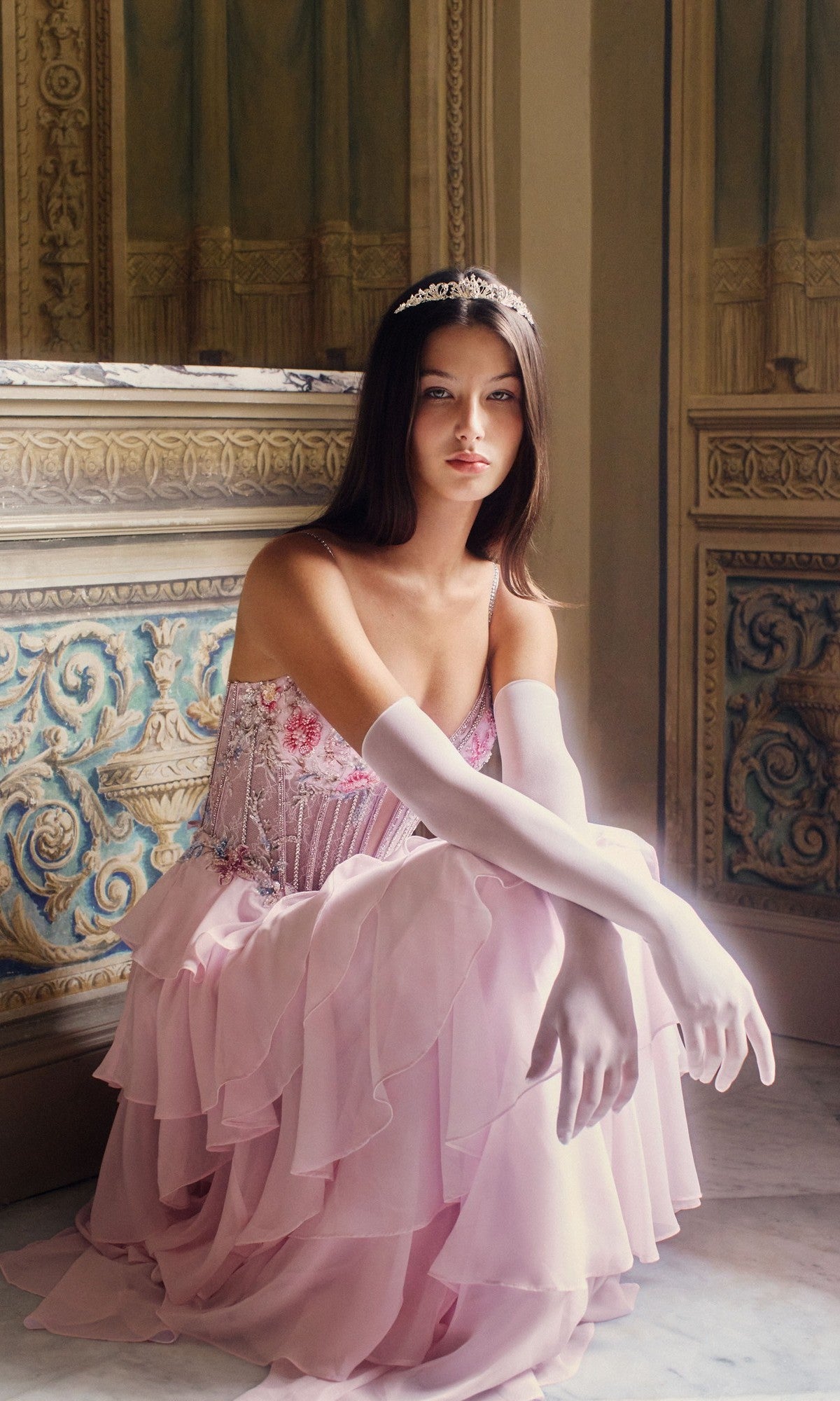 A woman in the Alyce 61993 long prom dress—a pink pastel gown with a chiffon tiered ruffle skirt—wears a tiara and white gloves as she sits on the floor of an ornate, decorative room.